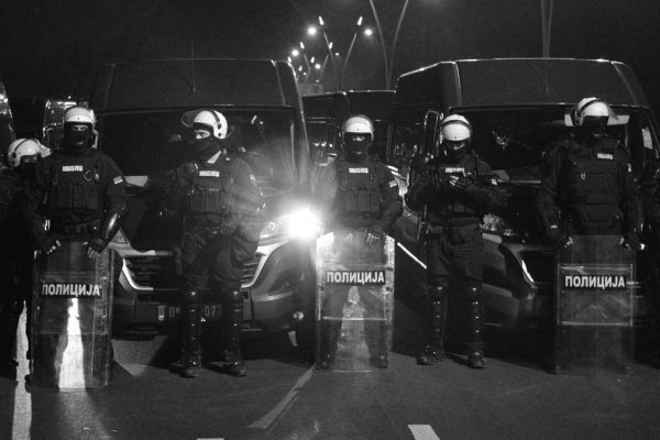 Riot police in protective gear and shields stand in line at night.